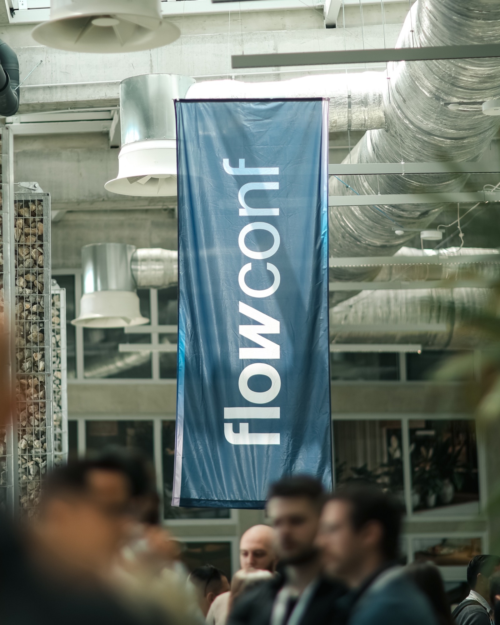 A flowConf banner hanging from the ceiling ducts of the hall, the crowd out of focus beneath it.