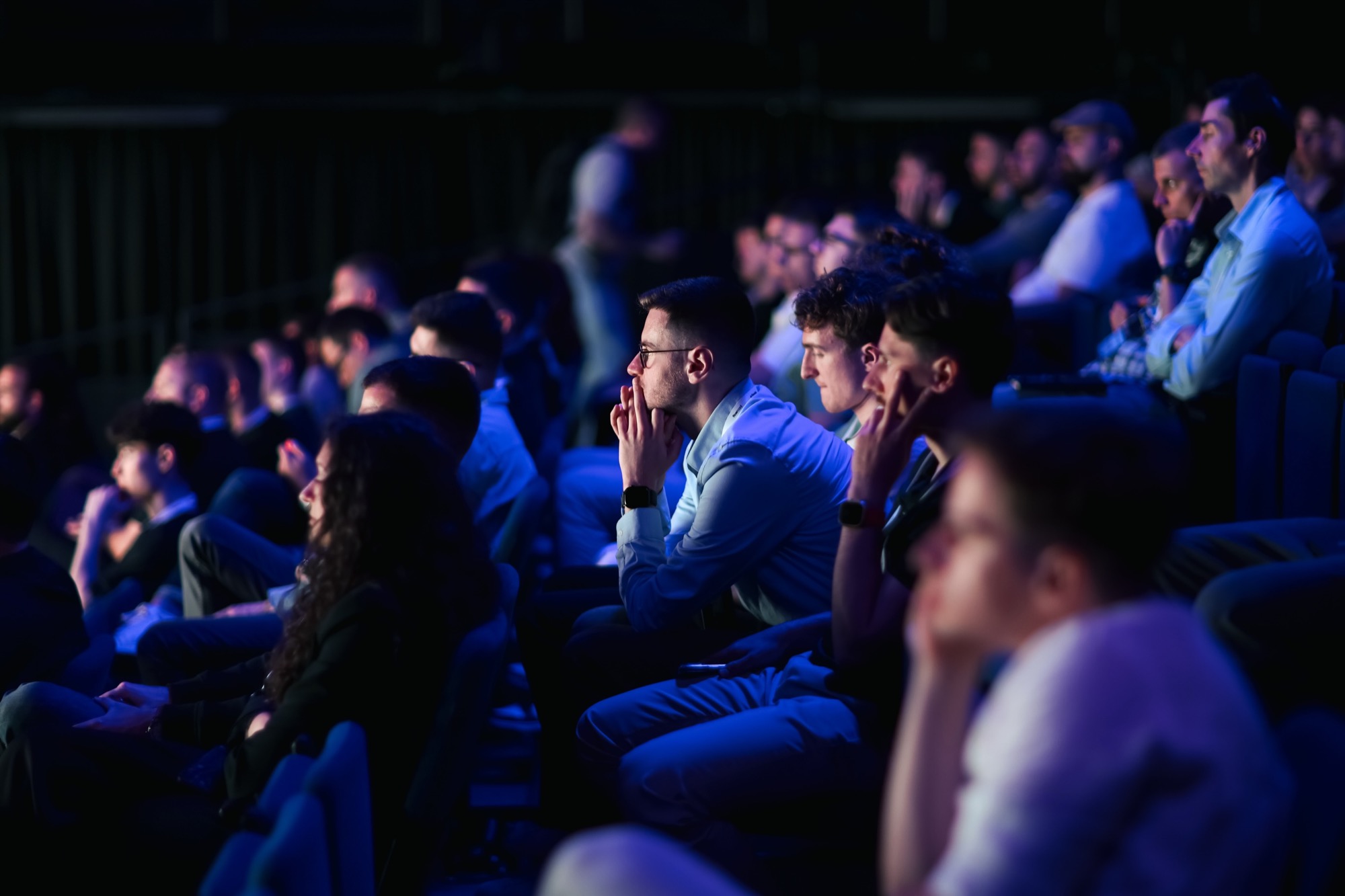 The auditorium in blue stage light, row after row of people watching a talk.