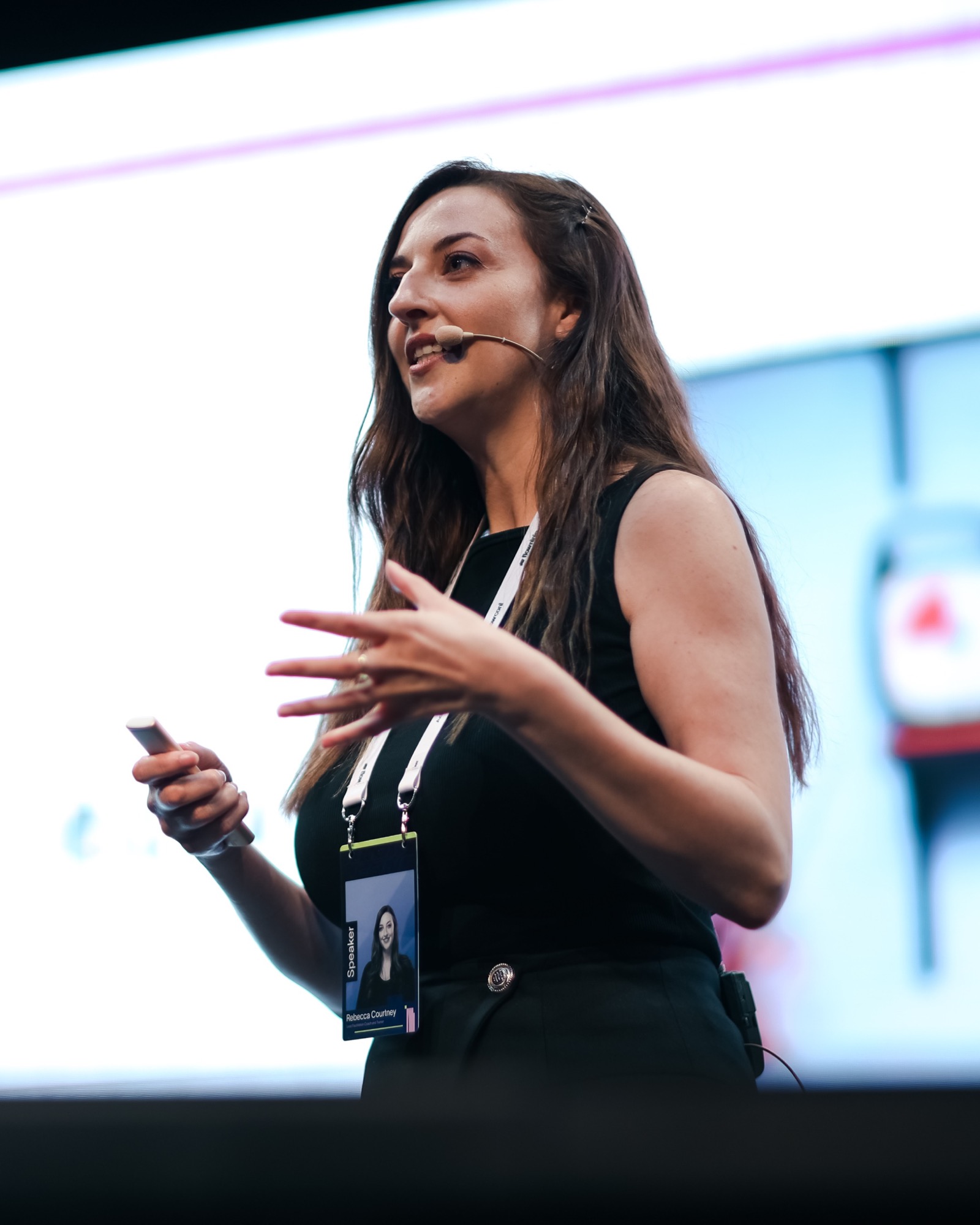 A speaker mid-sentence on stage, headset mic on and clicker in hand, against the glow of the screen behind her.