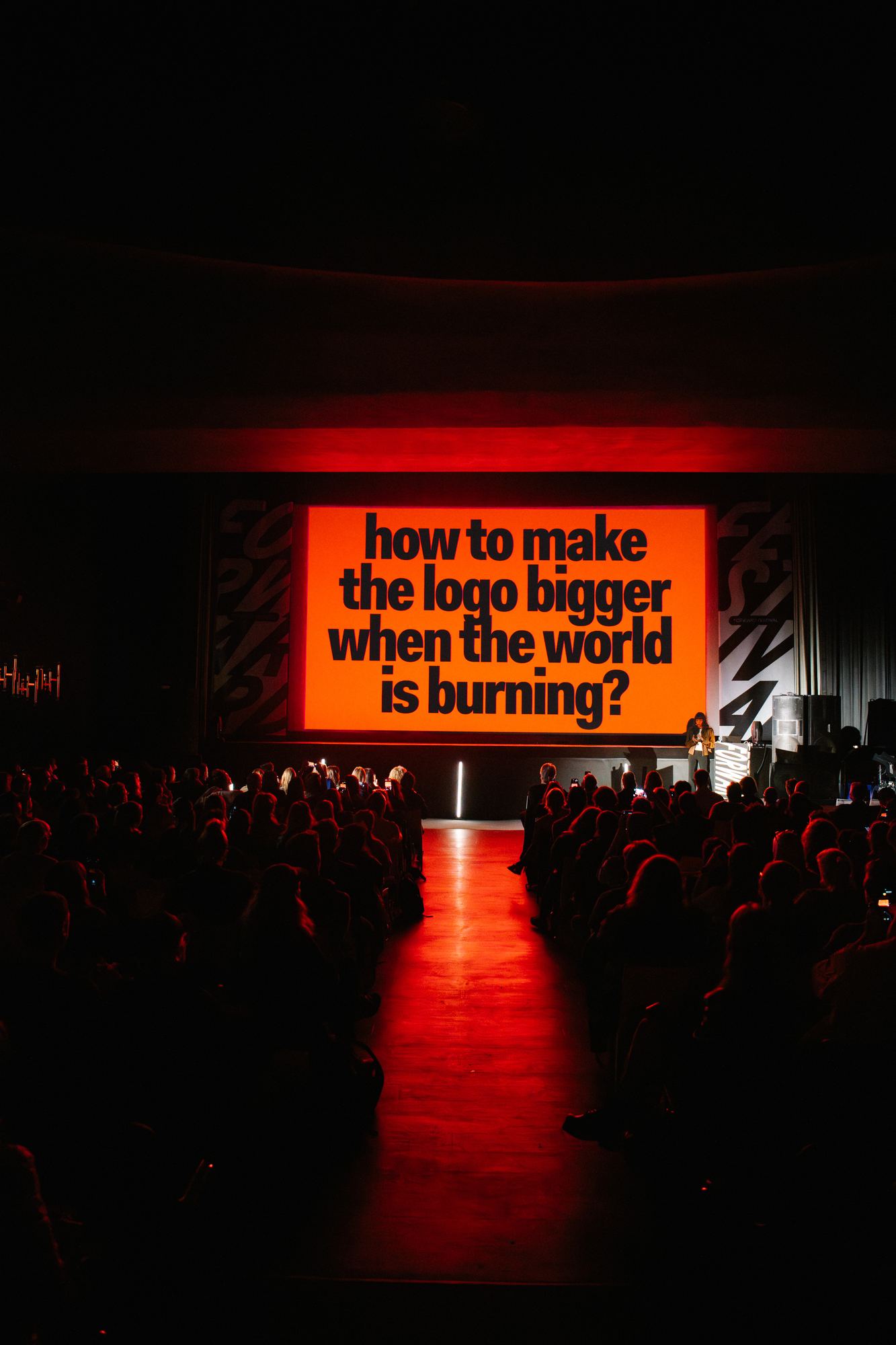 A full auditorium lit red by the screen, which asks: how to make the logo bigger when the world is burning?