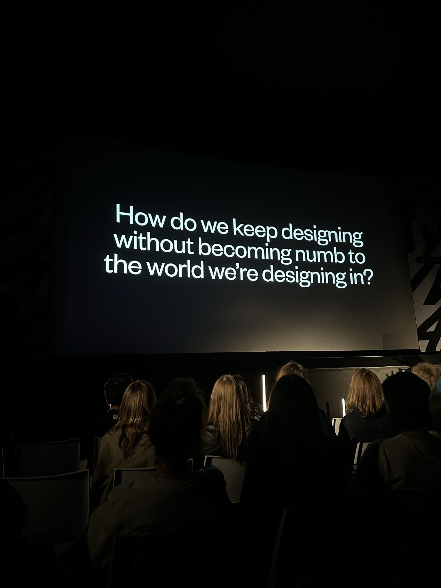 The auditorium seen from the audience, the screen asking how we keep designing without becoming numb to the world we're designing in.