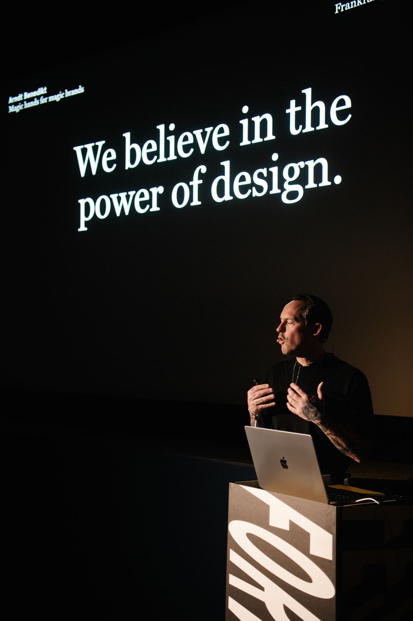 A speaker mid-sentence beside a slide reading "We believe in the power of design".