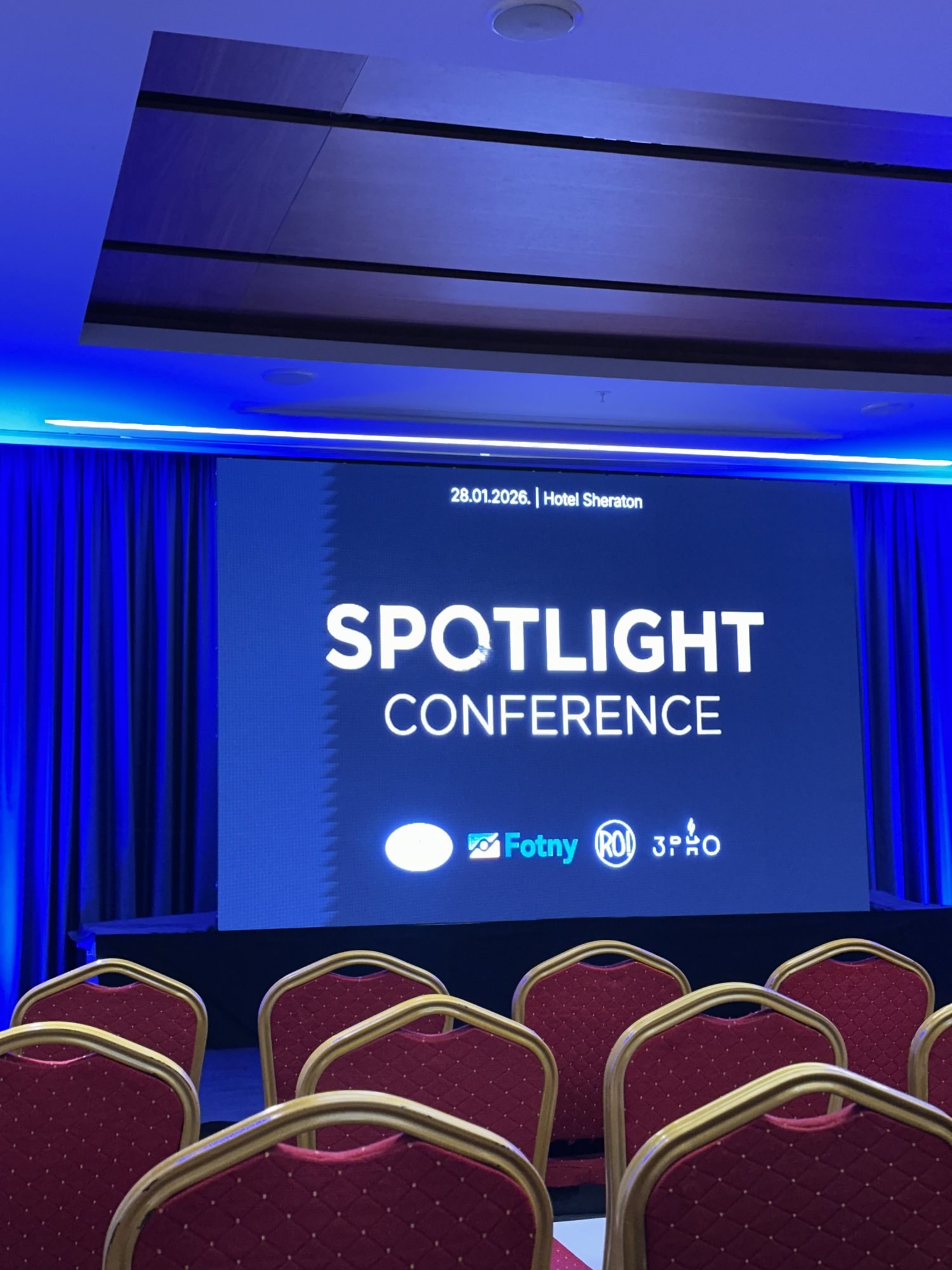 The stage screen in the Sheraton hall under blue light, the Spotlight Conference title slide up with "28.01.2026. | Hotel Sheraton" and the sponsor logos below it.
